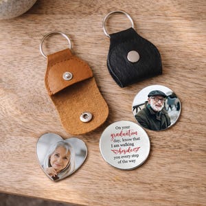 May include: A collection of personalized keychains and photo keepsakes. Includes a heart-shaped photo charm, two round photo charms, and two leather keychain holders in brown and black. One charm has the text: "On your graduation day, know that I am walking beside you every step of the way."