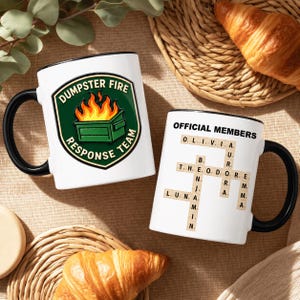 May include: Two white ceramic mugs with black handles. One mug features a green dumpster fire graphic with the text DUMPSTER FIRE RESPONSE TEAM. The other mug displays the text OFFICIAL MEMBERS above a crossword puzzle of names including Olivia, Aurora, Theodore, Benjamin, Luna, and Emma.
