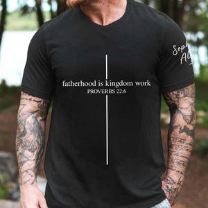 May include: Black crew neck t-shirt with the text "fatherhood is kingdom work" and "PROVERBS 22:6" in white. A vertical white line is centered on the shirt. The shirt has short sleeves.