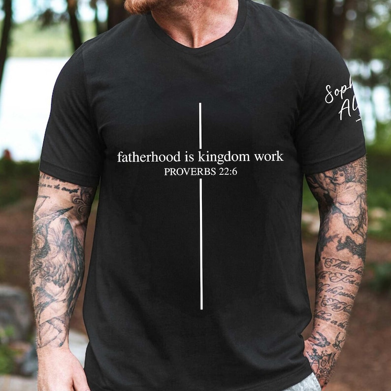 May include: Black crew neck t-shirt with the text "fatherhood is kingdom work" and "PROVERBS 22:6" in white. A vertical white line is centered on the shirt. The shirt has short sleeves.