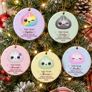 May include: Five round ceramic ornaments in pastel colours, each featuring a cute cartoon dumpling character with a face and the text: THE YEAR [Name] DID A LOT OF SQUISHY HUNTING 2026. The ornaments hang by gold strings against a decorated pine tree.