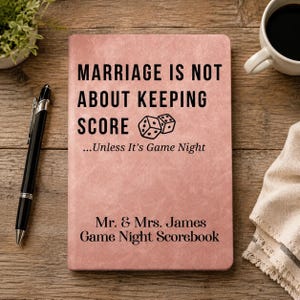 Peut inclure: Un carnet rose poudré avec un texte noir indiquant : MARRIAGE IS NOT ABOUT KEEPING SCORE ...Unless It’s Game Night, accompagné d'un dessin de deux dés, et le texte : Mr. & Mrs. James Game Night Scorebook.