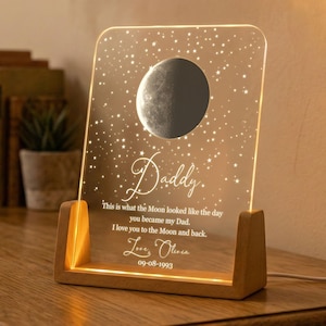 May include: A clear acrylic night light with a moon and star design, set in a wooden base. The light is engraved with the word "Daddy" and a personalized message. The date "09-08-1993" is also included.