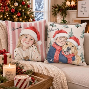 May include: Two square throw pillows featuring custom illustrations. The left pillow has pink and white stripes and a girl wearing a Santa hat with the name Hannah. The right pillow has a cream background with trees, a boy and dog with the name William.