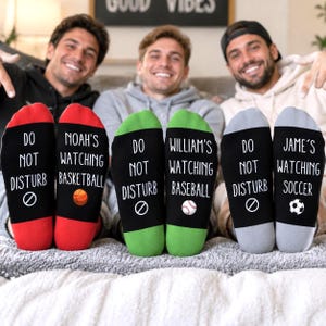 May include: Three pairs of personalised socks with black soles featuring the text DO NOT DISTURB and a prohibition symbol. The red pair says NOAH'S WATCHING BASKETBALL, the green pair says WILLIAM'S WATCHING BASEBALL, and the grey pair says JAME'S WATCHING SOCCER.