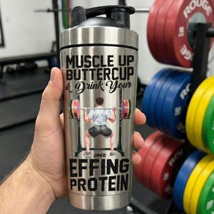 Puede incluir: Una botella mezcladora de acero inoxidable con tapa negra. La botella presenta el texto "MUSCLE UP BUTTERCUP & Drink Your EFFING PROTEIN" y una ilustraci&oacute;n de un levantador de pesas. La botella est&aacute; sujeta en una mano.