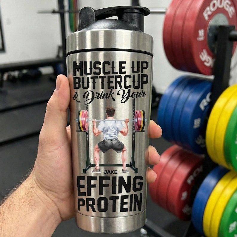 Puede incluir: Una botella mezcladora de acero inoxidable con tapa negra. La botella presenta el texto "MUSCLE UP BUTTERCUP & Drink Your EFFING PROTEIN" y una ilustraci&oacute;n de un levantador de pesas. La botella est&aacute; sujeta en una mano.