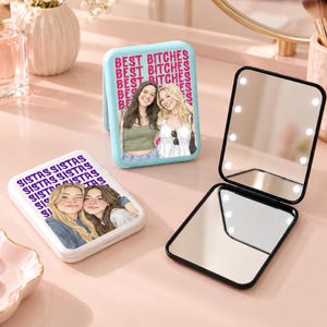 May include: Three rectangular compact mirrors. One light blue mirror features a graphic of two people and the text BEST BITCHES. One white mirror shows an illustration of two people and the text SISTAS. One black mirror is open, revealing LED lights.