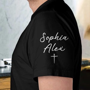 May include: Black t-shirt with white script text that reads "Sophia Alex" and a small cross below. The shirt is made of a soft material and is likely a personalized item.
