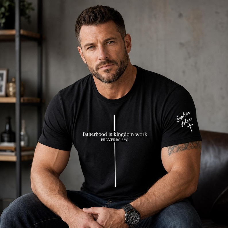 May include: Black t-shirt with the text "fatherhood is kingdom work" and "PROVERBS 22:6" in white. A white cross and the name "Sophia Alex" are on the right sleeve. The shirt is worn by a person.