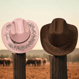 May include: A light pink cowboy hat and a dark brown cowboy hat, each featuring a braided band and a floral design engraved on the brim. The pink hat is personalised with the name Sophia, and the brown hat is personalised with the name Emily.