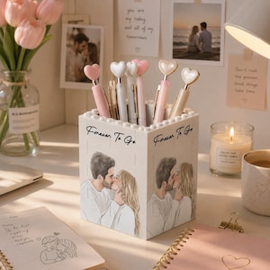 May include: A white square pen holder constructed from building blocks, featuring the text "Forever To Go" and a printed illustration of a couple kissing. The holder contains several pens topped with heart-shaped charms in shades of pink and white.