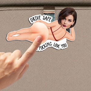 May include: A car visor clip featuring a cartoon illustration of a person in a red bikini with a photographic face cutout. The text reads DRIVE SAFE and I FUCKING LOVE YOU. The design includes a squishy, rounded element representing a body part.