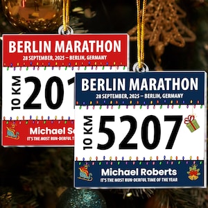 May include: Two Christmas tree decorations shaped like Berlin Marathon race bibs. One is red with the number 201, the other is navy blue with 5207. Both have the text "BERLIN MARATHON" and "10 KM" displayed.