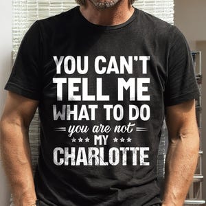May include: A black short-sleeved t-shirt featuring white distressed-style text that reads: YOU CAN'T TELL ME WHAT TO DO you are not MY CHARLOTTE. The design includes decorative horizontal lines and three small stars on each side of the word MY.