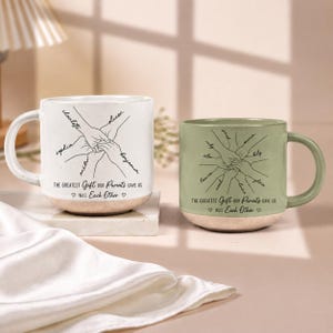 May include: Two ceramic mugs, one white and one sage green, each featuring a black line drawing of hands joined together with names written along the arms and the text, "The greatest gift our parents gave us was each other," with small hearts.