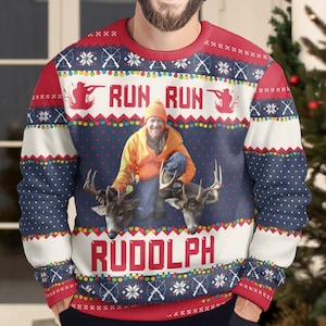 May include: A long-sleeved crewneck jumper featuring a dark blue base with white polka dots, red and white bands, and a festive holiday pattern. The centre displays a photo of a person in orange hunting gear with two deer, with the text RUN RUN RUDOLPH.