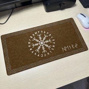 May include: A rectangular brown desk mat featuring a white Vegvisir symbol centred on the surface, surrounded by a decorative braided border. The background includes a subtle pattern of faint runic symbols, and the text NM+R is printed in white.