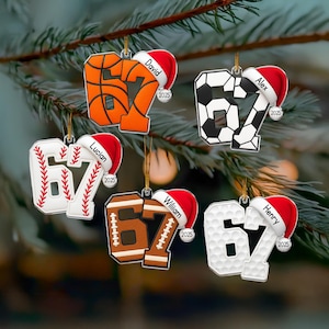 May include: A selection of Christmas tree decorations, each shaped as the number 67 in the style of different sports balls. Includes basketball, football, baseball, golf ball, and soccer ball designs, each with a Santa hat and a name.