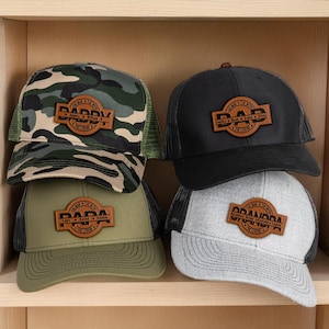 May include: Four trucker caps with leather patches. One has a camouflage pattern, another is black, a third is olive green, and the last is grey. Each patch reads "DAD" or "DADDY" with names below and "THE MAN THE MYTH THE LEGEND" around the edge.
