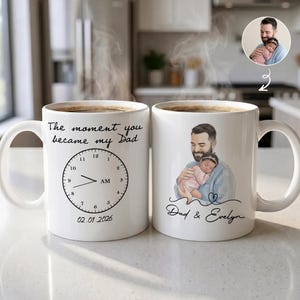 Pode incluir: Duas canecas de cer&acirc;mica branca com caf&eacute;. Uma caneca tem o texto "The moment you became my Dad" com um design de rel&oacute;gio e a data 02.01.2026. A outra caneca tem uma ilustra&ccedil;&atilde;o de um pai segurando um beb&ecirc; e o texto "Dad & Evelyn."
