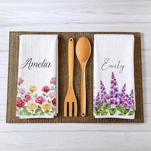 May include: Two white waffle-weave kitchen towels featuring floral prints and names. One towel displays the name Amelia with red, pink, and yellow roses. The other displays the name Emily with purple flowers. A wooden spoon and fork sit between them.