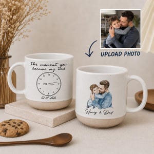 May include: Two white ceramic mugs with a beige base. One mug has a clock design and the text "The moment you became my Dad" and the date 02.01.2026. The other mug has a watercolor image of a father and child with the text "Henry & Dad".