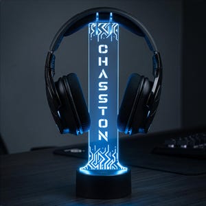May include: A black headset rests on a vertical, illuminated acrylic stand. The stand features a glowing blue light, a circuit board pattern, and the word CHASSTON printed vertically in white capital letters on a rectangular base.