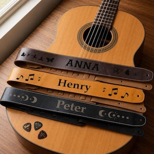 May include: Three leather guitar straps resting on an acoustic guitar. The top strap is dark brown with butterfly motifs and the name ANNA. The middle strap is tan with musical notes and the name Henry. The bottom strap is black with moon motifs and the name Peter. Three guitar picks are included.