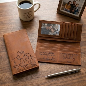 May include: A brown leather wallet with a photo window and multiple card slots. The wallet is open, revealing a picture and engraved text. A line drawing of a father and child is on the front. A coffee mug and pen are also on the wooden surface.
