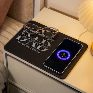 May include: A black wireless charging pad with the text "DAD" and "WE LOVE YOU EVERY DAY". Gold-rimmed glasses and a smartphone charging at 5% are on the pad. The pad is on a white nightstand.