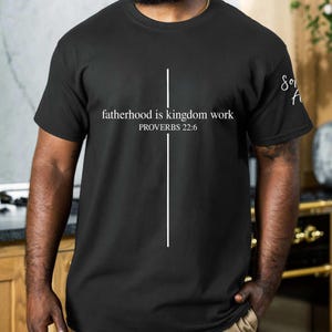 May include: Black t-shirt with the text "fatherhood is kingdom work" and "PROVERBS 22:6" in white. A vertical white line intersects the text. The sleeve has the text "Son of A" in white. The shirt is a crew neck.