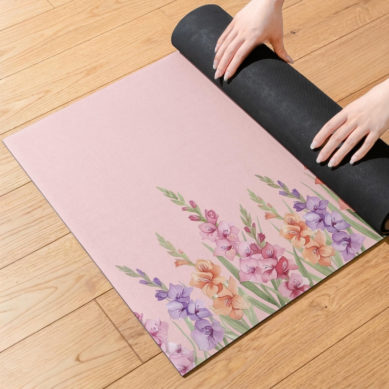 May include: A rectangular yoga mat with a soft pink background featuring a watercolor-style floral print of gladiolus flowers in shades of purple, orange, and pink. The mat has a black underside and is partially rolled up on one side.
