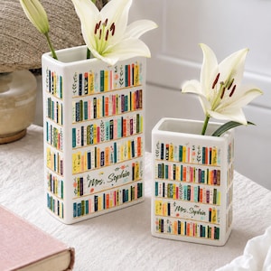 May include: Two rectangular white ceramic vases featuring a colorful graphic print of stacked books on shelves. Each vase displays the text Mrs. Sophia in cursive script and holds a single white lily flower with green stems and dark stamens.