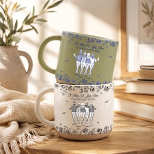 Könnte beinhalten: Zwei vertikal gestapelte Keramikbecher. Der obere Becher ist salbeigrün mit blauen Blumenmustern und einer Grafik von vier Freunden mit dem Text It Takes A Long Time TO GROW OLD FRIENDS. Der untere Becher ist weiß mit schwarzen Blumenmustern.