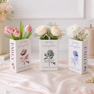 May include: Three rectangular white ceramic vases shaped like books. Each vase features the text THE NEXT chapter, a floral illustration, a name, and the phrase every chapter brings new growth. The vases contain pink tulips, white roses, and white chrysanthemums.