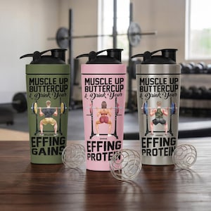 Puede incluir: Tres botellas de coctelera de acero inoxidable con tapas negras. Cada botella presenta un color diferente: verde oliva, rosa y plateado. Las botellas tienen el texto "MUSCLE UP BUTTERCUP & Drink Your" y un gr&aacute;fico de levantamiento de pesas.