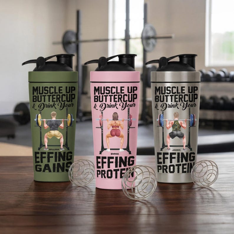 Puede incluir: Tres botellas de coctelera de acero inoxidable con tapas negras. Cada botella presenta un color diferente: verde oliva, rosa y plateado. Las botellas tienen el texto "MUSCLE UP BUTTERCUP & Drink Your" y un gr&aacute;fico de levantamiento de pesas.