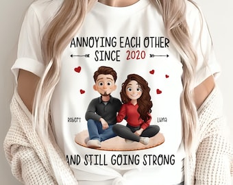 May include: A white t-shirt featuring a cartoon illustration of a couple sitting together. The text reads "ANNOYING EACH OTHER SINCE 2020" and "AND STILL GOING STRONG" with the names "ROBERT" and "LUNA" printed near the characters, surrounded by red hearts.