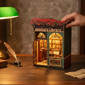 May include: A miniature library model with the text "Jessica's Library" on the front. The model is green and red with an open door revealing a detailed interior. A green desk lamp, a book, and a teacup are also on the desk.