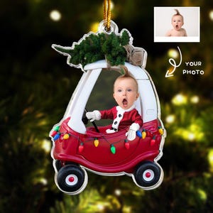 May include: A personalized Christmas ornament featuring a baby in a red toy car, wearing a Santa suit. The car is decorated with colorful lights and a small Christmas tree. The ornament includes a space for a photo.