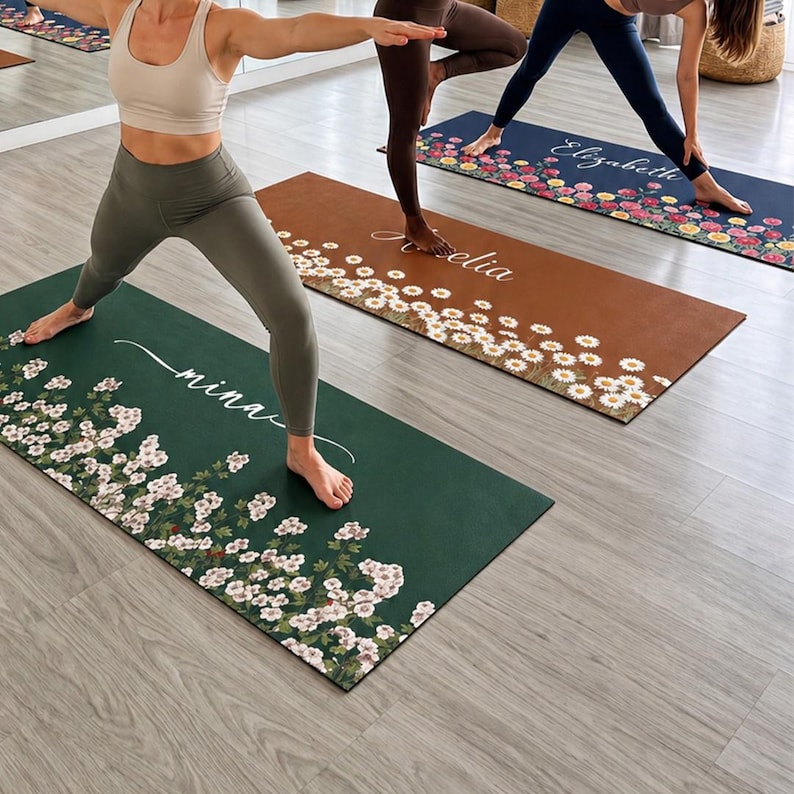 May include: Three rectangular yoga mats featuring floral patterns and personalized names in white script. One mat is dark green with white flowers and the name 'mina', one is brown with white daisies and 'Amelia', and one is navy with colorful flowers and 'Elizabeth'.