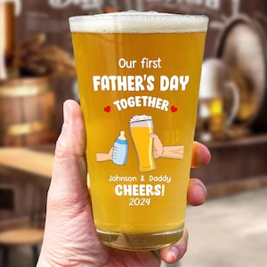 May include: A clear glass beer mug with a yellow liquid inside. The mug has a design that says "Our first Father's Day Together" with a baby bottle and a beer glass. The text also says "Johnson & Daddy Cheers! 2024".