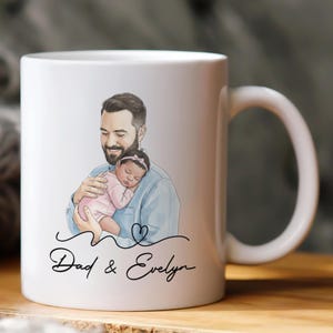 Pode incluir: Caneca de cer&acirc;mica branca com uma ilustra&ccedil;&atilde;o em aquarela de um pai segurando um beb&ecirc; dormindo. A caneca tem o texto "Dad & Evelyn" em uma fonte manuscrita, com um design de cora&ccedil;&atilde;o. A al&ccedil;a &eacute; branca.