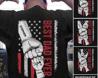 May include: A black t-shirt featuring a graphic of a large fist bumping a smaller fist over a stylized American flag background. The text BEST DAD EVER is printed vertically in red. The fists include customizable name labels like STEPHEN and NOAH.