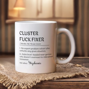 May include: White ceramic mug with the text "CLUSTER FUCK FIXER" in black, along with a definition. The mug also includes the name "Stephanie". The mug is on a burlap surface.