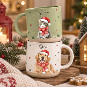 May include: Two ceramic mugs stacked vertically. The top sage green mug features a tabby cat wearing a red Santa hat and scarf with the name Luna. The bottom cream mug features a golden retriever wearing a red Santa hat and scarf with the name Oreo.