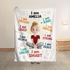 May include: A white blanket with a cartoon illustration of a girl, with the name "AMELIA" above her. The blanket has positive affirmations like "I am KIND", "I am BRAVE", and "I am SMART" in various colours and fonts, with a drawing of a basketball.