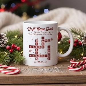 May include: White ceramic mug with the text "Best Team Ever" and a crossword puzzle design featuring names and the year 2025. The mug has a curved handle and is decorated with festive elements.