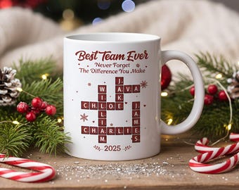 May include: White ceramic mug with the text "Best Team Ever" and a crossword puzzle design featuring names and the year 2025. The mug has a curved handle and is decorated with festive elements.